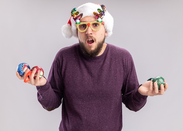 Foto gratuita giovane uomo in maglione viola e cappello da babbo natale indossando occhiali divertenti tenendo le palle di natale guardando la fotocamera confusa in piedi su sfondo bianco