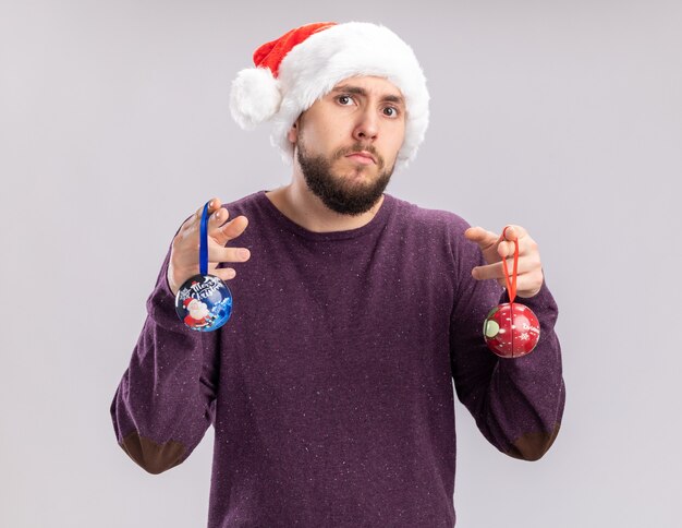 보라색 스웨터와 산타 모자에 젊은 남자가 카메라를보고 크리스마스 공을 들고 재미있는 안경을 쓰고 흰색 배경 위에 서 혼란