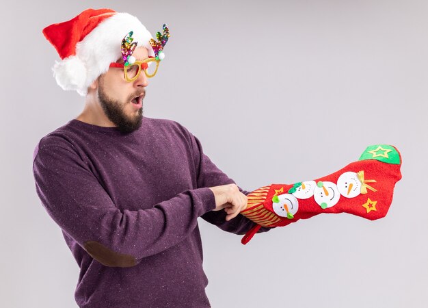 Young man in purple sweater and santa hat wearing funny glasses holding a christmas stocking looking at it amazed and surprised standing over white background