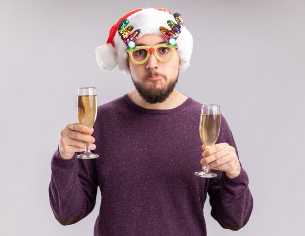 紫色のセーターとサンタの帽子をかぶった若い男は、白い背景の上に立っているシャンパンの2つのグラスを保持している面白い眼鏡をかけています。