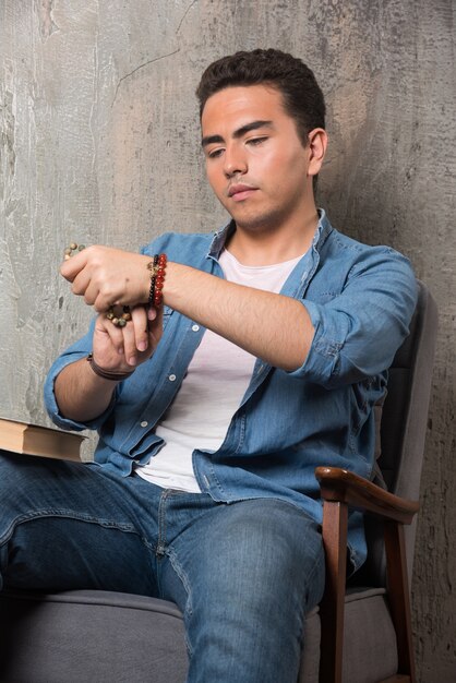 Young man putting on bracelet and sitting on chair with book on marble background. High quality photo