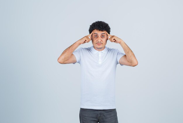 Young man putting fingers on temples in white t-shirt and jeans and looking serious , front view.