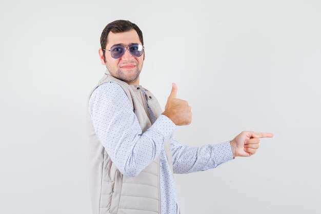 Молодой человек надевает очки, указывая вправо указательным пальцем и показывает большой палец в бежевой куртке и кепке и выглядит оптимистично. передний план.