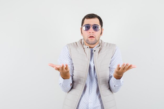 Young man putting on glasses while raising hands as receiving something in beige jacket and cap and looking surprised. front view.