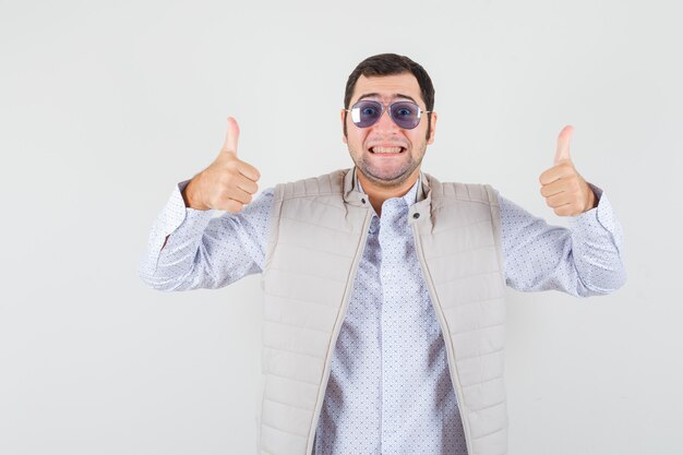 Young man putting on glasses while showing double thumbs up in beige jacket and cap and looking optimistic , front view.