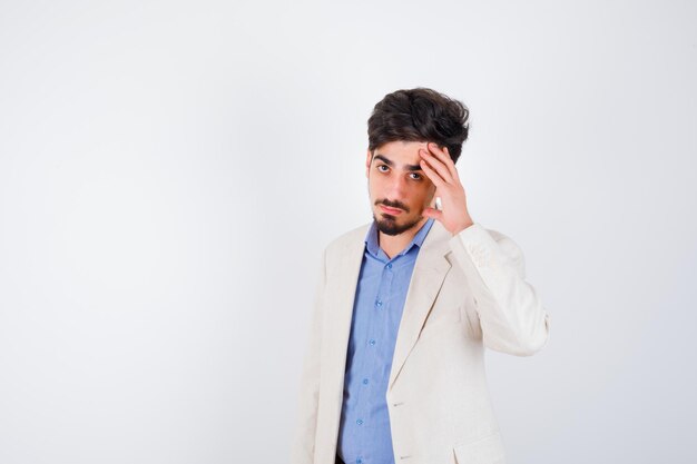 Young man putting hand to face, posing at front in blue t-shirt and white suit jacket and looking serious