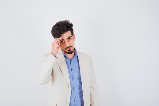 Young man putting hand to face, posing at front in blue t-shirt and white suit jacket and looking serious