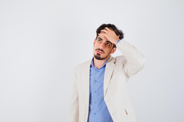 Young man putting hand on forehead, looking away in blue t-shirt and white suit jacket and looking pensive