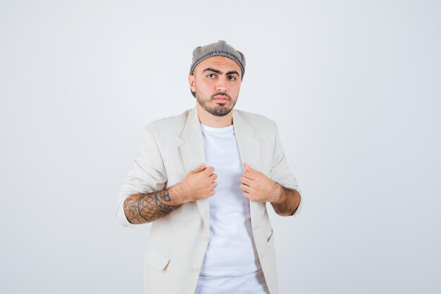 Young man putting hands on jacket in white t-shirt, jacket and gray cap and looking serious. front view.