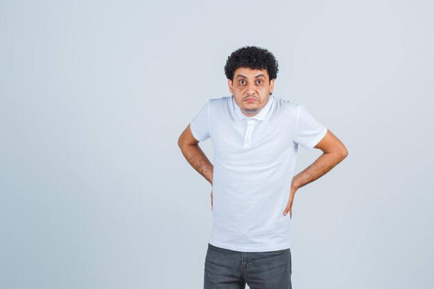 Young man putting hands on waist in white t-shirt and jeans and looking serious. front view.