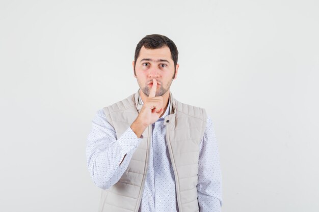 Young man putting index finger on mouth and showing silence gesture in beige jacket and looking serious. front view.