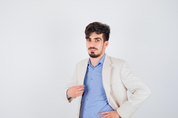 Young man putting one hand on jacket, another hand on waist in blue shirt and white suit jacket and looking confident