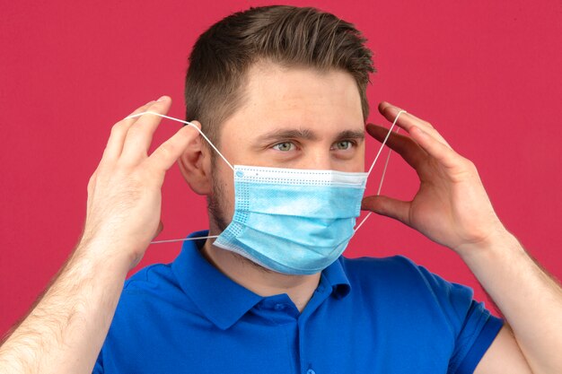 Молодой человек надевает защитную медицинскую маску на лицо над изолированной розовой стеной