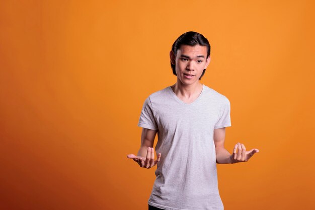 Young man questioning, shrugging shoulders with unsure facial expression. Puzzled asian teenager standing with hands spread wide, pensive person portrait, medium shot on orange background