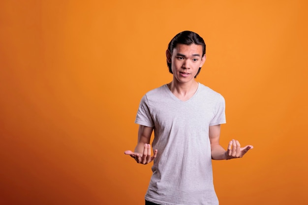 Молодой человек допрашивает, пожимая плечами с неуверенным выражением лица. Озадаченный азиатский подросток, стоящий с широко раскинутыми руками, портрет задумчивого человека, средний снимок на оранжевом фоне