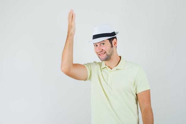 Молодой человек поднимает руку в футболке, шляпе и выглядит веселым.