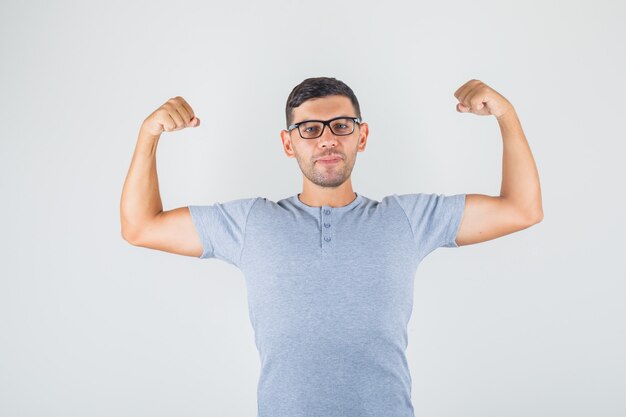 Young man raising arms and showing biceps in grey t-shirt, glasses front view.