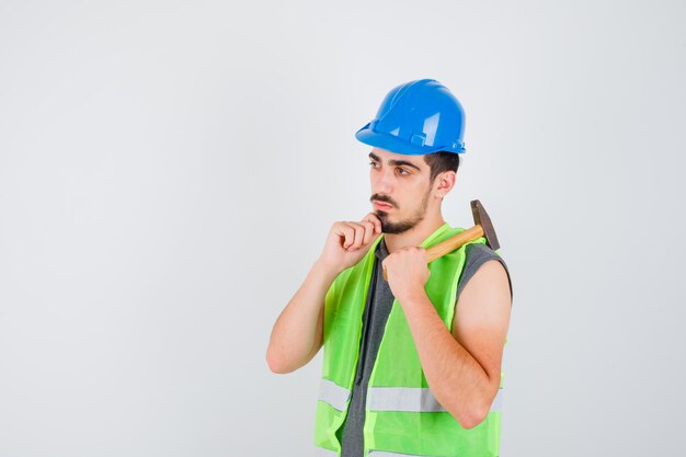 Young man raising axe over shoulder and leaning chin on hand in construction uniform and looking pensive