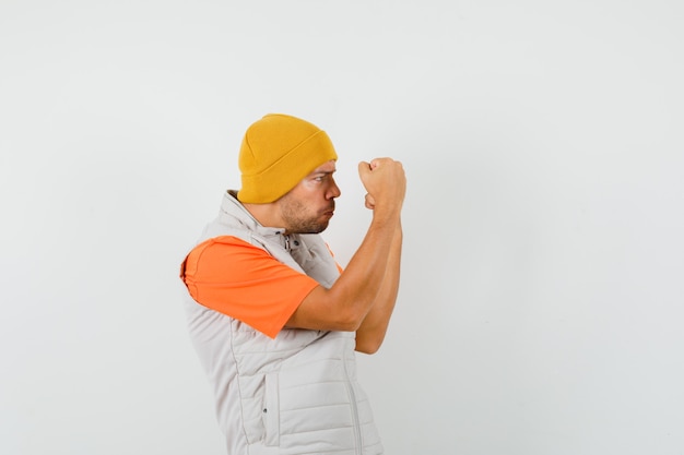 Young man raising clenched fists in t-shirt, jacket, hat and looking strong