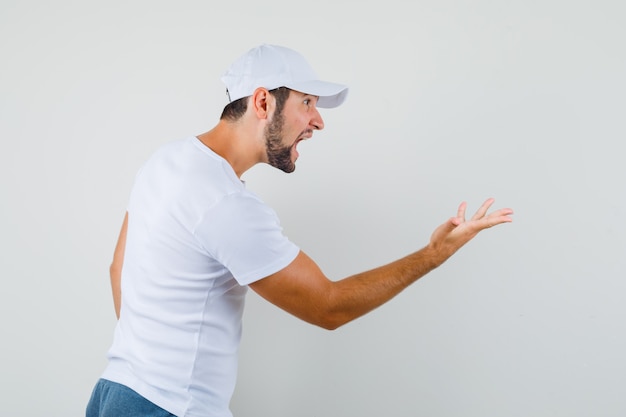 Young man raising hand in questioning gesture aside while shouting in white t-shirt,cap and looking angry .