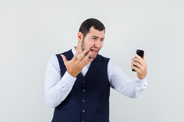 Young man raising hand while using video chat in shirt and vest and looking aggressive