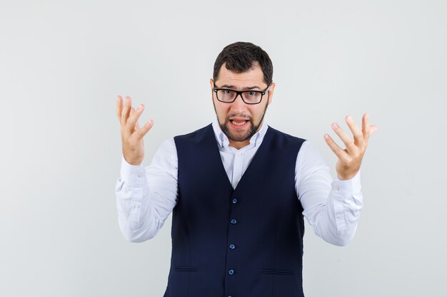 Young man raising hands in aggressive manner in shirt and vest and looking nervous