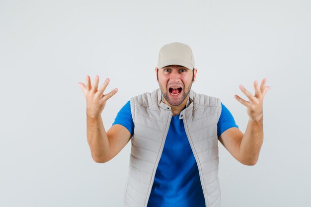 Young man raising hands in aggressive manner while shouting in t-shirt, jacket, cap front view.