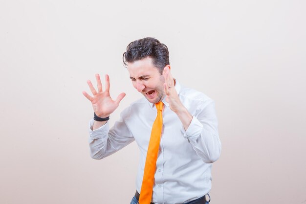 Young man raising hands in aggressive manner in white shirt, tie and looking agitated