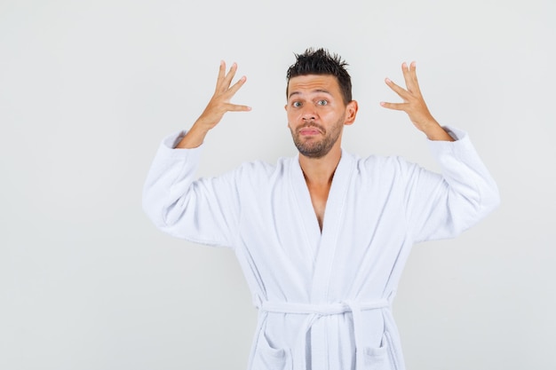 Young man raising hands in helpless manner in white bathrobe front view.
