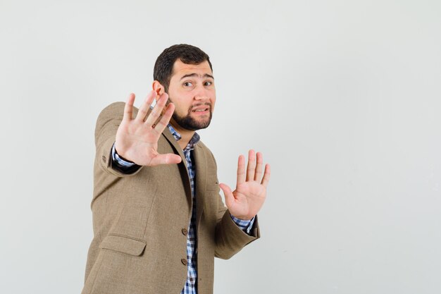 Young man raising hands in protective manner in shirt, jacket and looking scared.