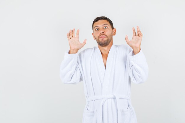 Young man raising hands in protective manner in white bathrobe and looking scared. front view.