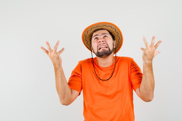 Young man raising hands in puzzled gesture in orange t-shirt, hat and looking mournful. front view.