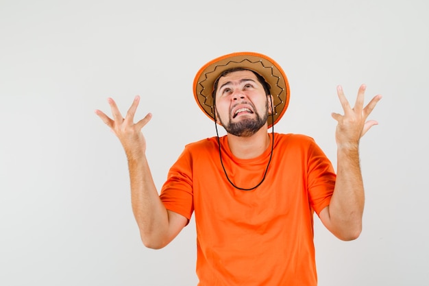 オレンジ色のTシャツ、帽子で困惑したジェスチャーで手を上げて、悲しそうに見える若い男。正面図。