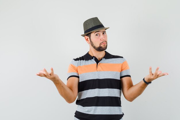 Young man raising hands in puzzled gesture in t-shirt, hat and looking doubtful.