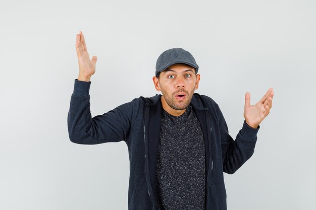 Young man raising hands in puzzled gesture in t-shirt, jacket, cap