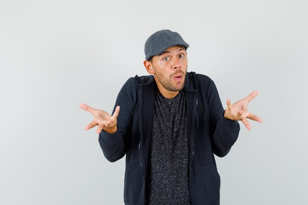 Young man raising hands in puzzled gesture in t-shirt, jacket, cap