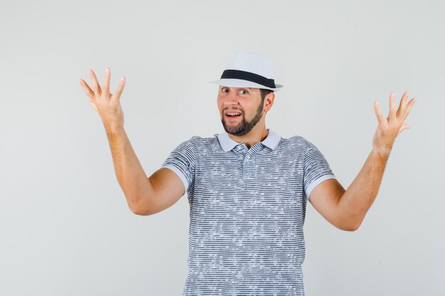Young man raising hands in questioning gesture in white t-shirt,hat and looking amazed , front view.