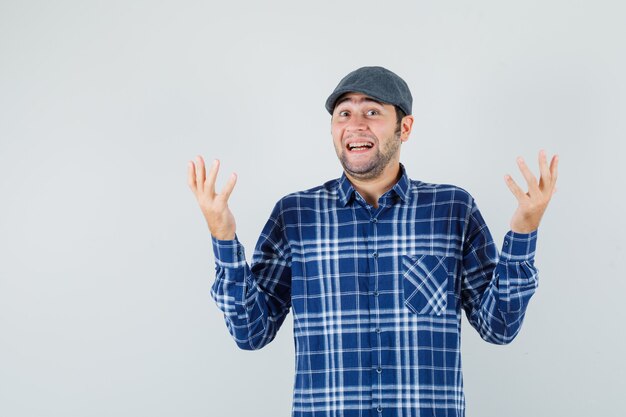 Young man raising hands in shirt, cap and looking merry , front view.