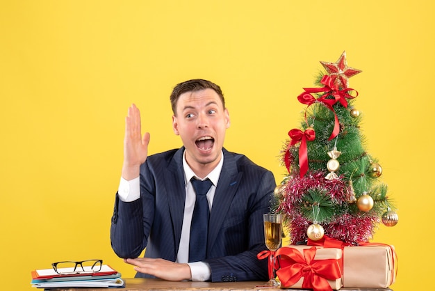 young man raising his hand sitting at the table near xmas tree and presents on yellow