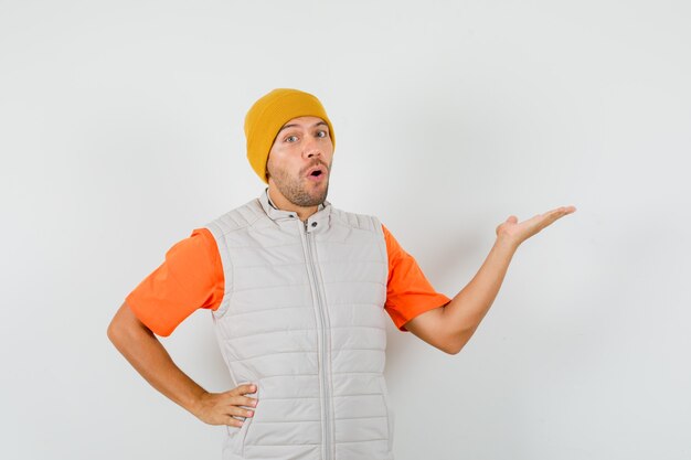 Young man raising palm as if holding or showing something in t-shirt, jacket, hat and looking amazed