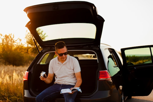 Young man reading a book and eating a chocolate on the car trunk