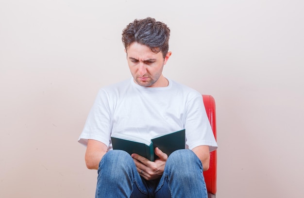 Giovane che legge un libro mentre è seduto su una sedia in maglietta, jeans e sembra perplesso