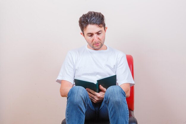 Young man reading book while sitting on chair in t-shirt, jeans