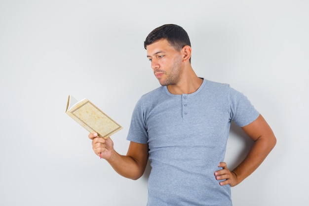 Free photo young man reading book with hand on waist in grey t-shirt and looking careful.