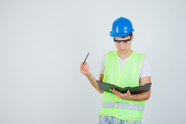 Young man reading notes in file folder while holding pen in construction uniform and looking focused