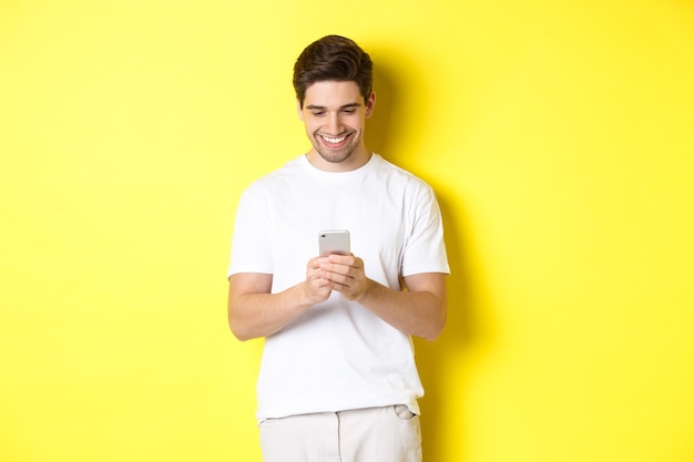 Молодой человек читает текстовое сообщение на смартфоне, смотрит на экран мобильного телефона и улыбается, стоя в белой футболке на желтом фоне