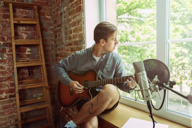 Young man recording music video blog, home lesson or song, playing guitar or making broadcast internet tutorial while sitting in loft workplace or at home