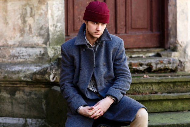 Young man in red hat and grey coat sits on ruined footsteps 