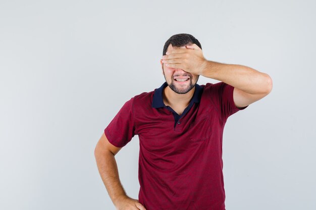 Young man in red t-shirt covering eyes with hand and looking excited , front view.