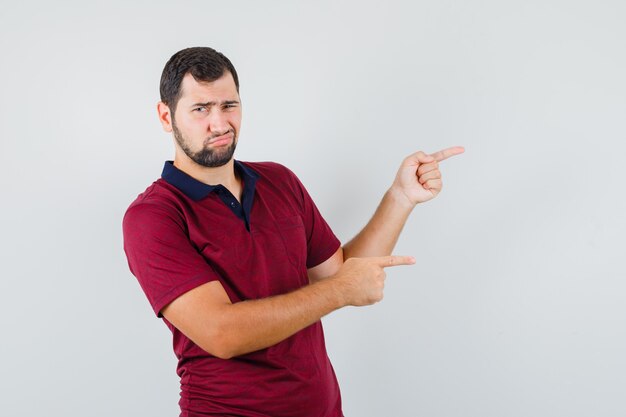 Young man in red t-shirt pointing to side and looking dissatisfied , front view.